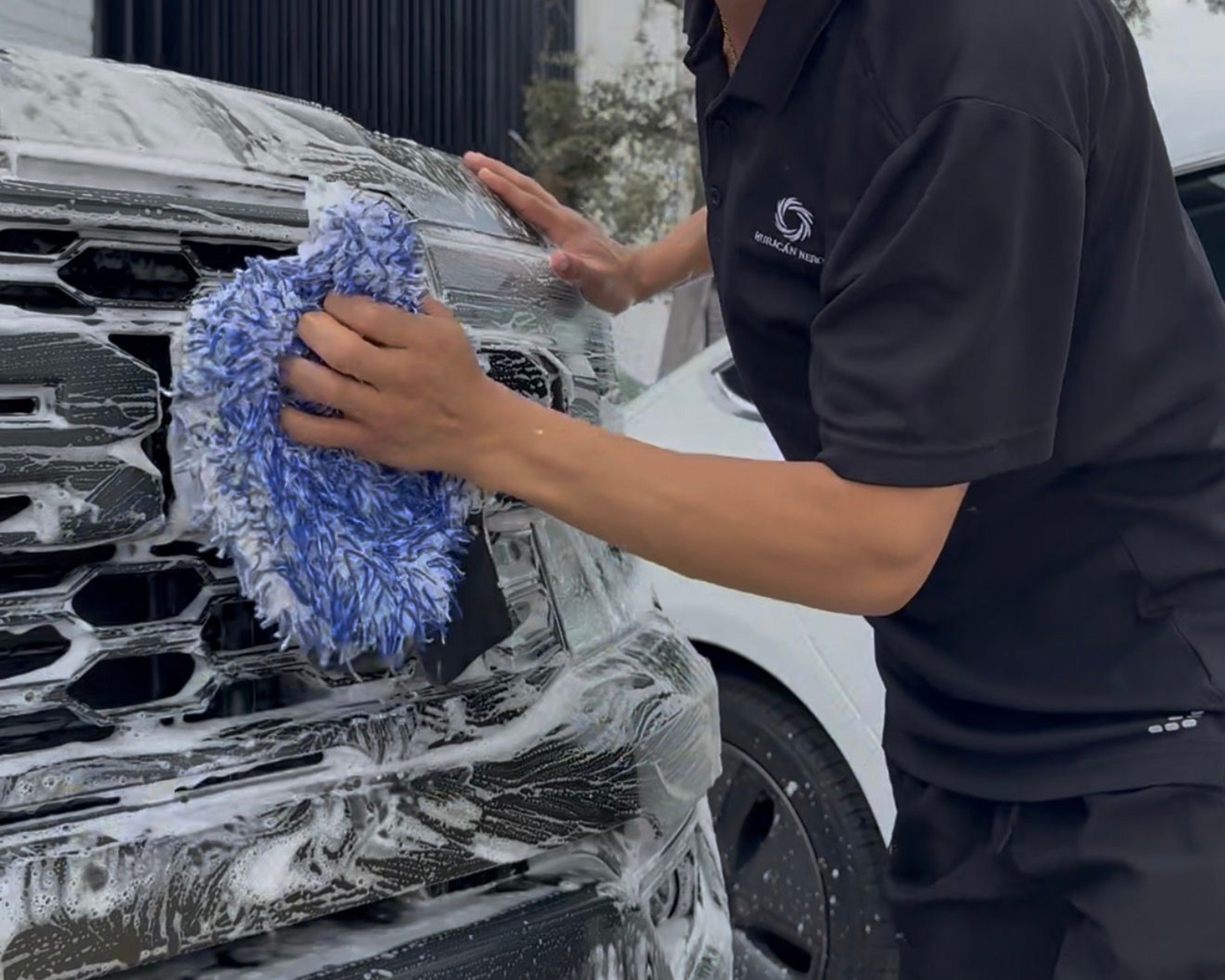 A person with curly hair wearing a black polo shirt washes a car grille covered in soap suds using a blue mitt.