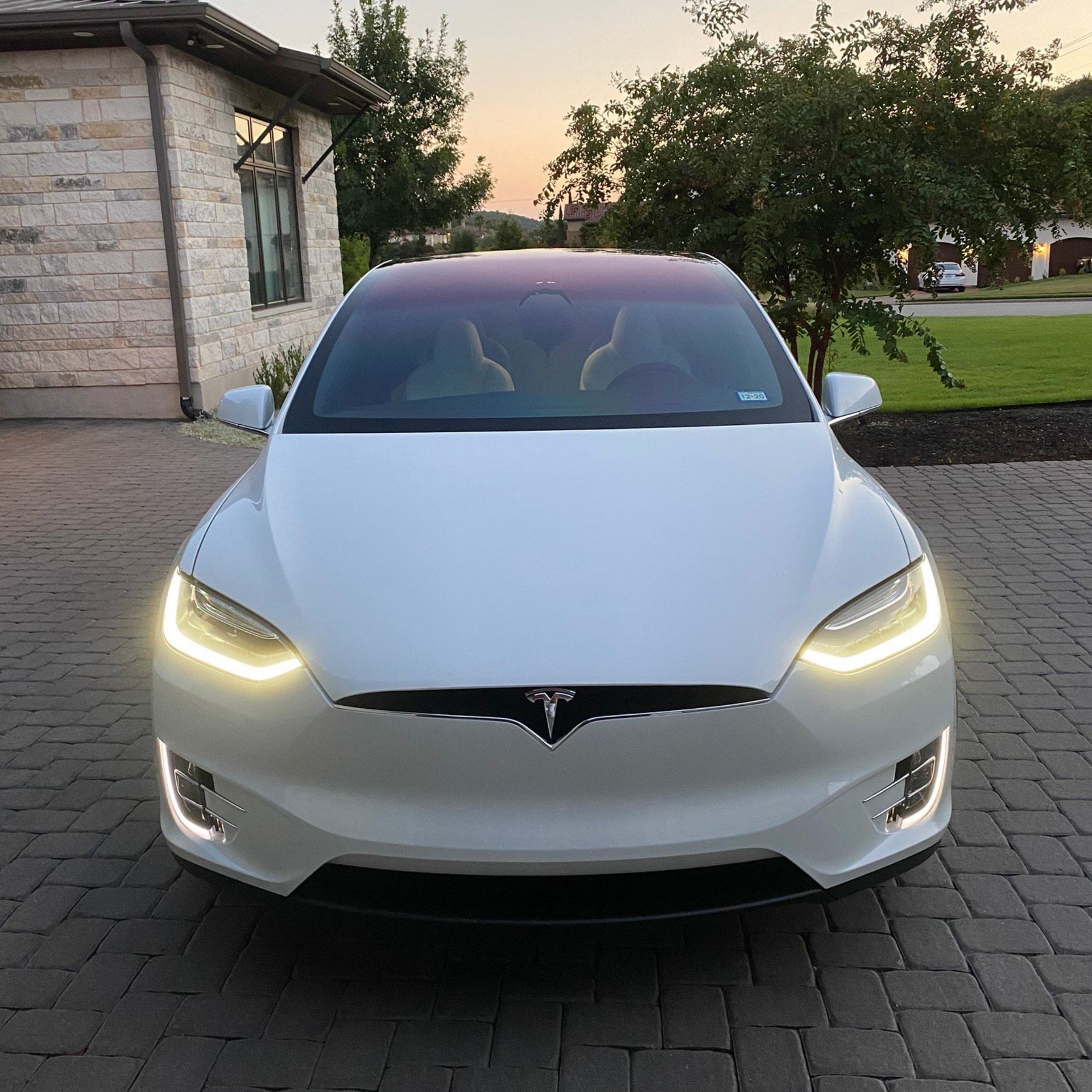 White Tesla Model X parked on a brick driveway, lights on, in front of a house.