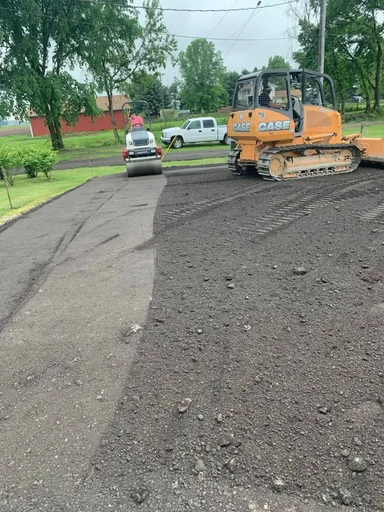 Road resurfacing with a yellow bulldozer spreading gravel beside a car on a tree-lined street.