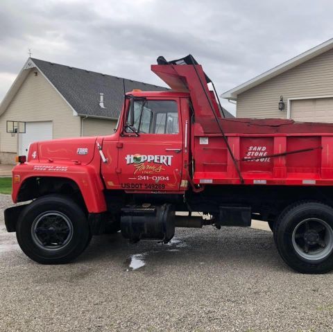 Red utility truck with raised dump bed parked in a residential driveway under cloudy skies
