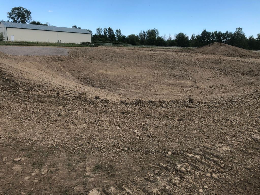 Dry gravel pit with a sloped dirt bank, trees in the background, and a white building at left.