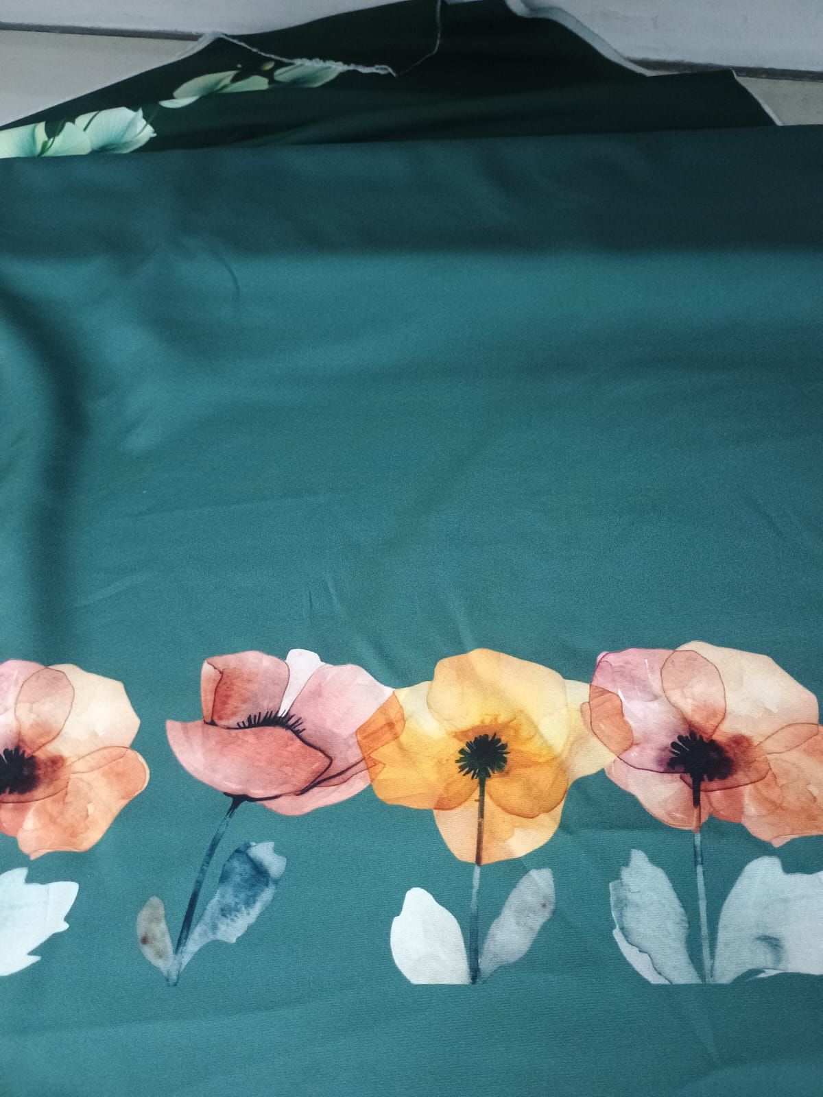 Una hilera de flores de amapola estilo acuarela en tonos melocotón y amarillo sobre un fondo de tela verde azulado intenso.