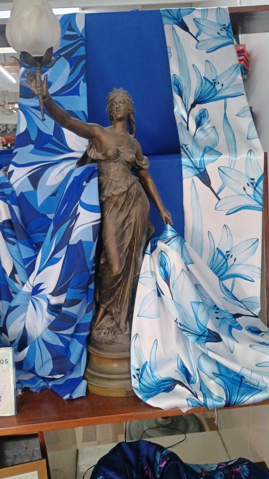 Una estatua clásica en tonos bronce que sostiene una pequeña lámpara, enmarcada por una tela floral azul