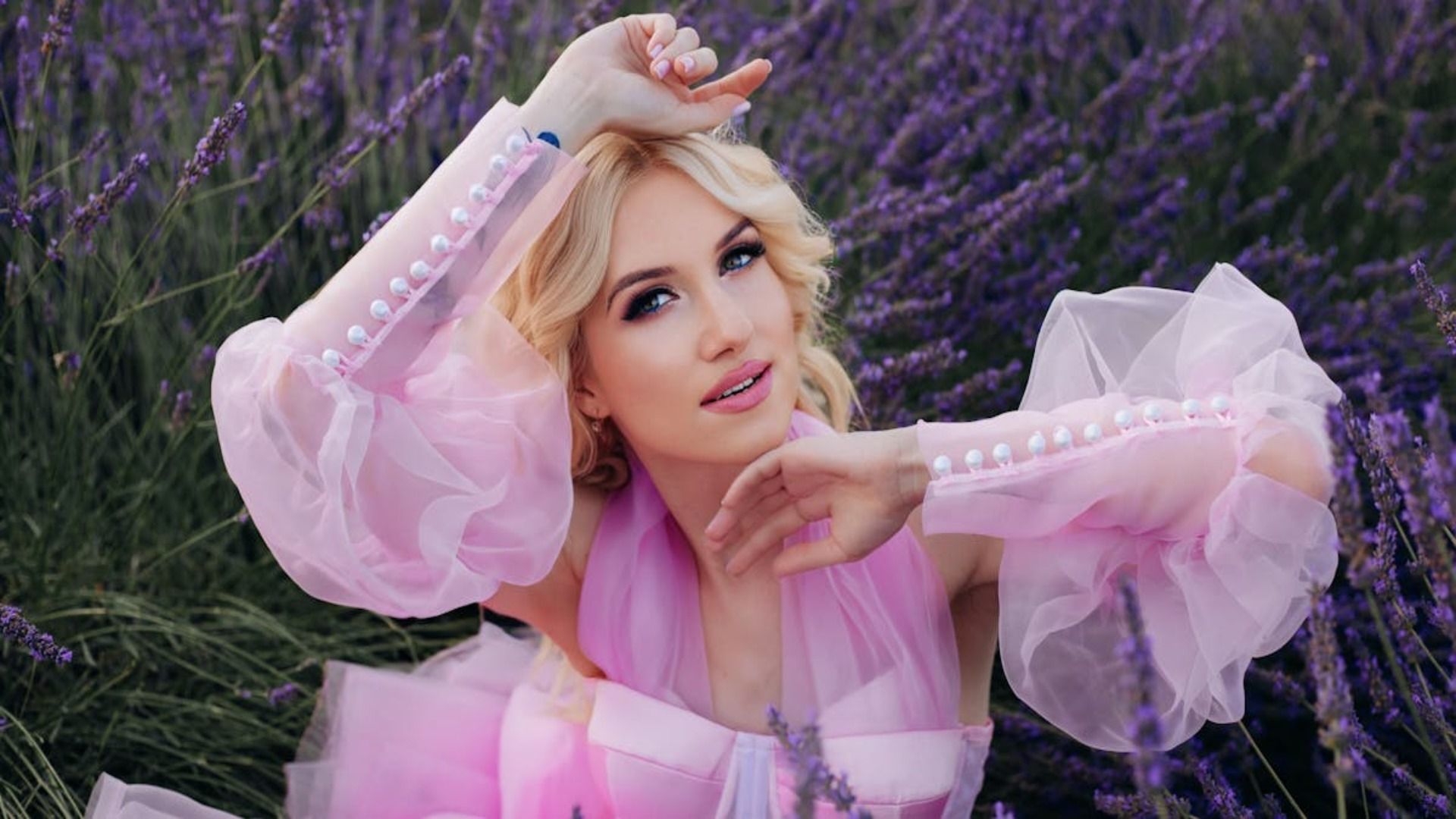 Una persona de cabello rubio con un vestido rosa transparente posa con los brazos levantados