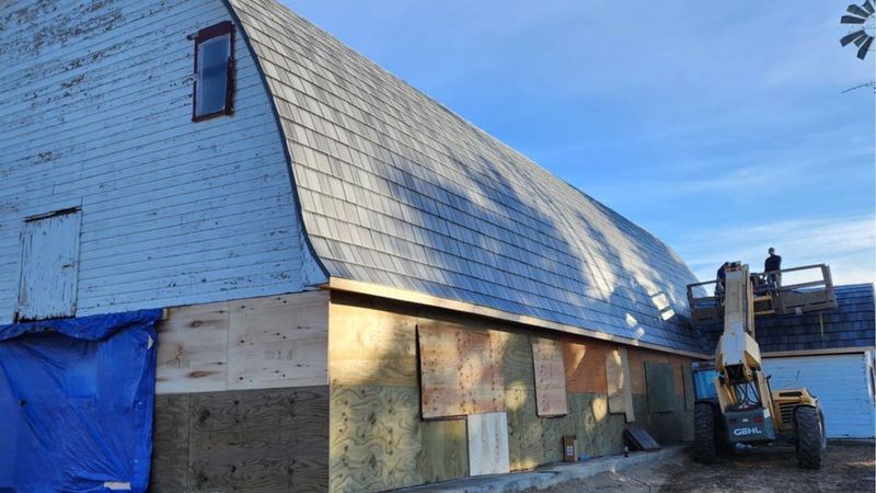 A blue, gambrel-roof barn undergoing repairs with plywood wall panels and a person on a lift working on the roof.