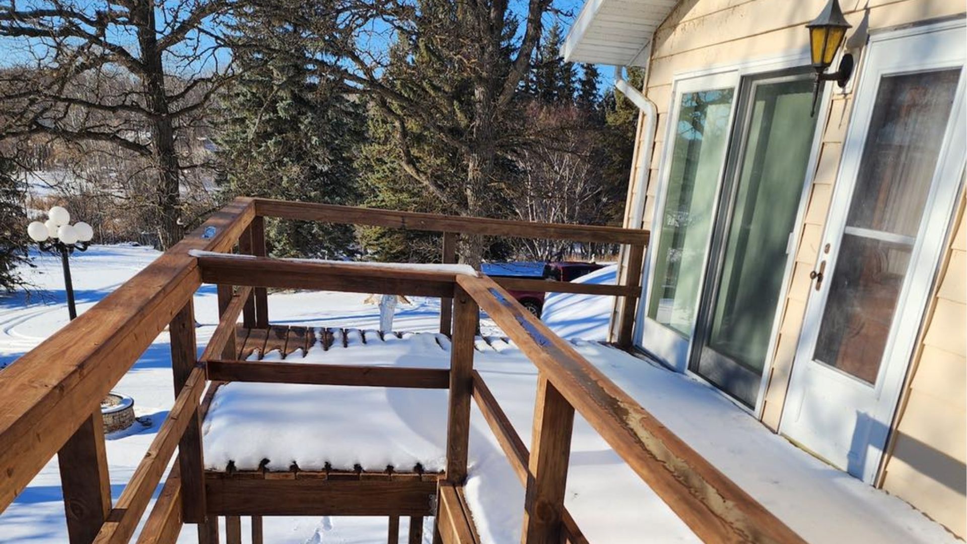 A snowy deck with wooden railings leads to glass patio doors on the side of a light-colored house.