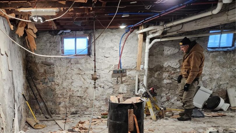 A person in a brown jacket stands in an unfinished, debris-filled basement with stone walls, exposed beams, and pipes.