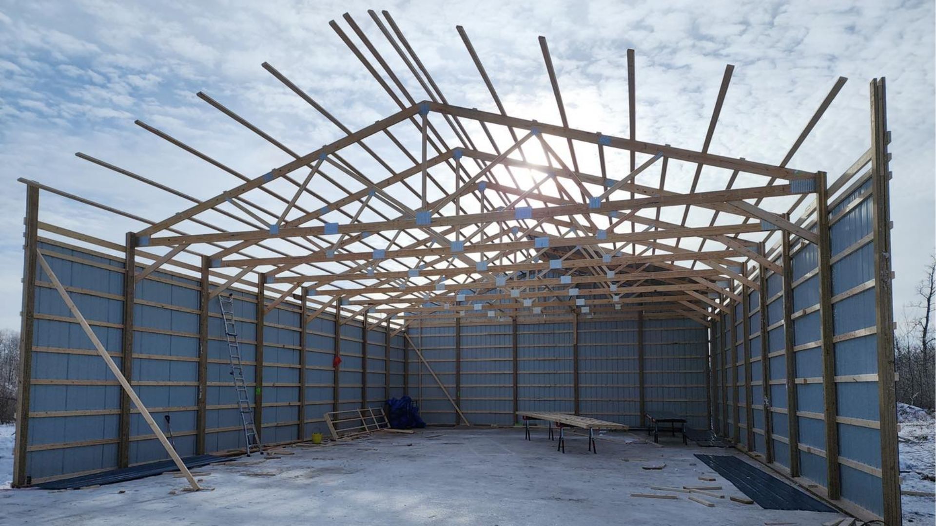 An open-frame construction of a pole barn featuring metal siding and an exposed wooden truss roof under a bright sky.