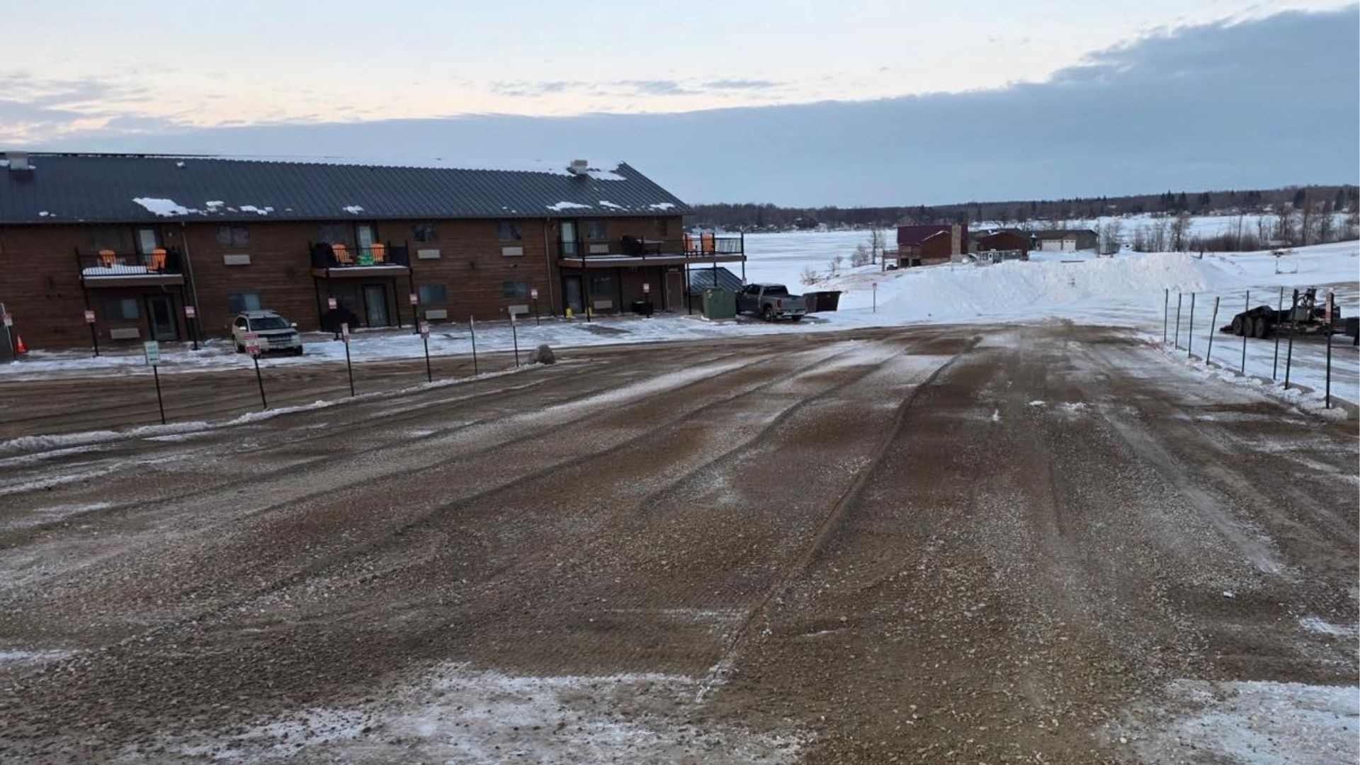 A gravel parking lot leads toward a long, brick building under a cloudy sky, with snow covering the surrounding ground.