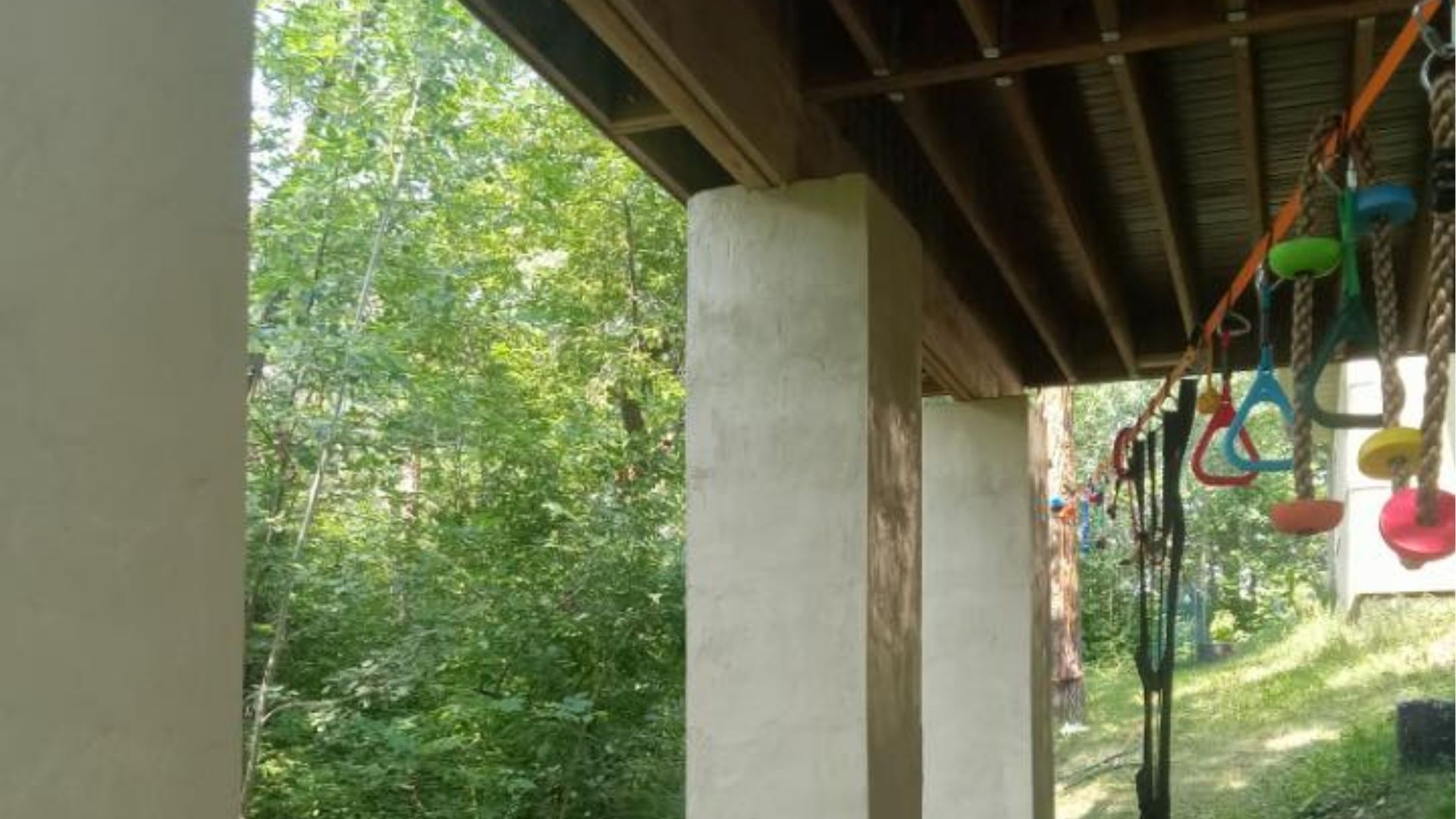 Outdoor play area with colorful hanging gymnastics rings and ropes attached under a wooden porch deck near a forest.