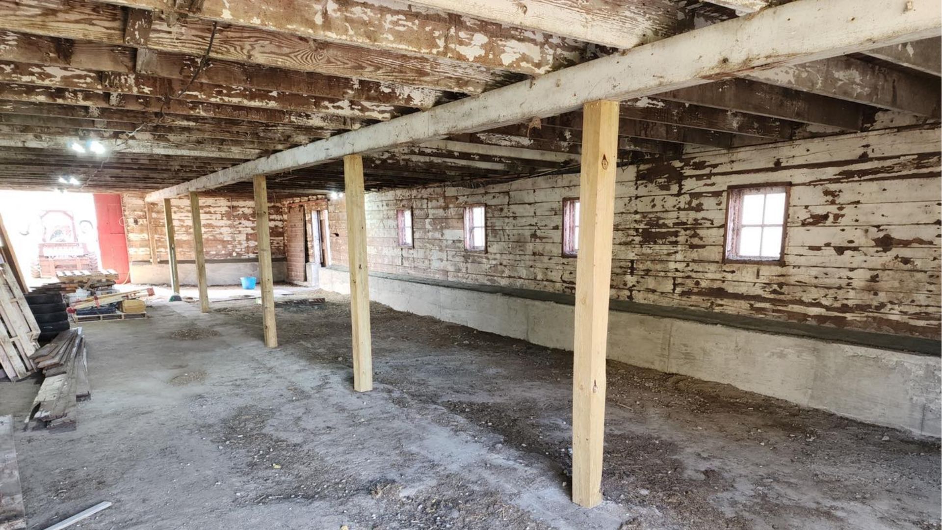 A wide-angle view of a rustic, unfinished basement with wooden support beams, dirt floors, and white-washed stone walls.