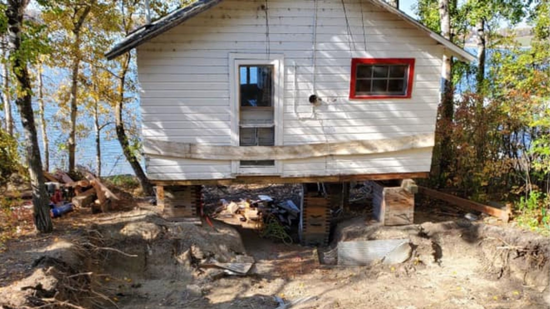 A white cottage-style structure elevated on temporary wooden piers over an excavated sandy ground in a forest setting.
