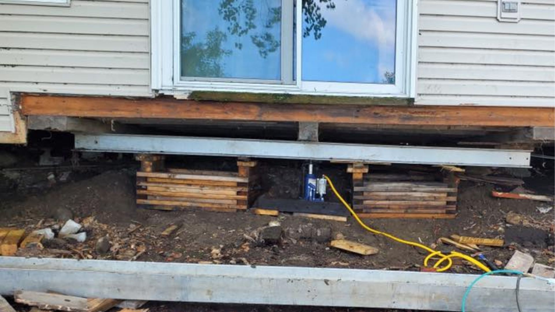 A mobile home exterior raised on two wooden cribbing stacks and a hydraulic jack, resting on dirt ground.