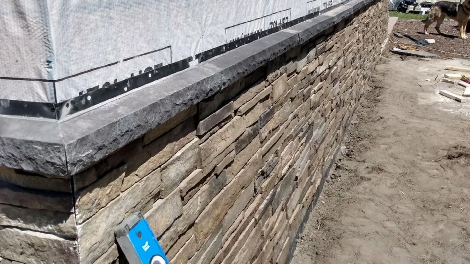 A stone veneer wall under construction with a dark gray stone cap and a blue level leaning against the corner.