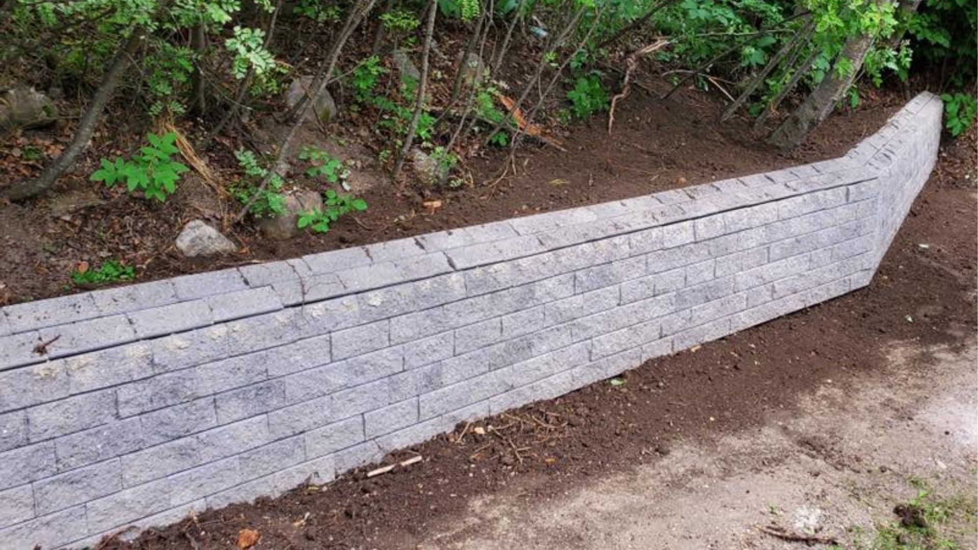 A newly constructed gray stone retaining wall curves along a sloped area of dark earth and wooded greenery.