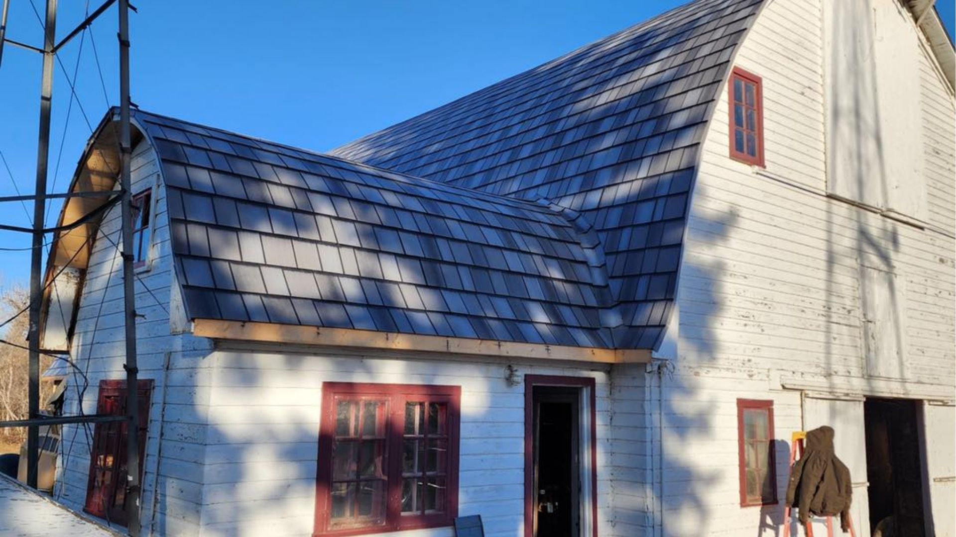 A white, two-story barn with a gambrel roof and dark shingles, featuring red window frames and a person standing outside.