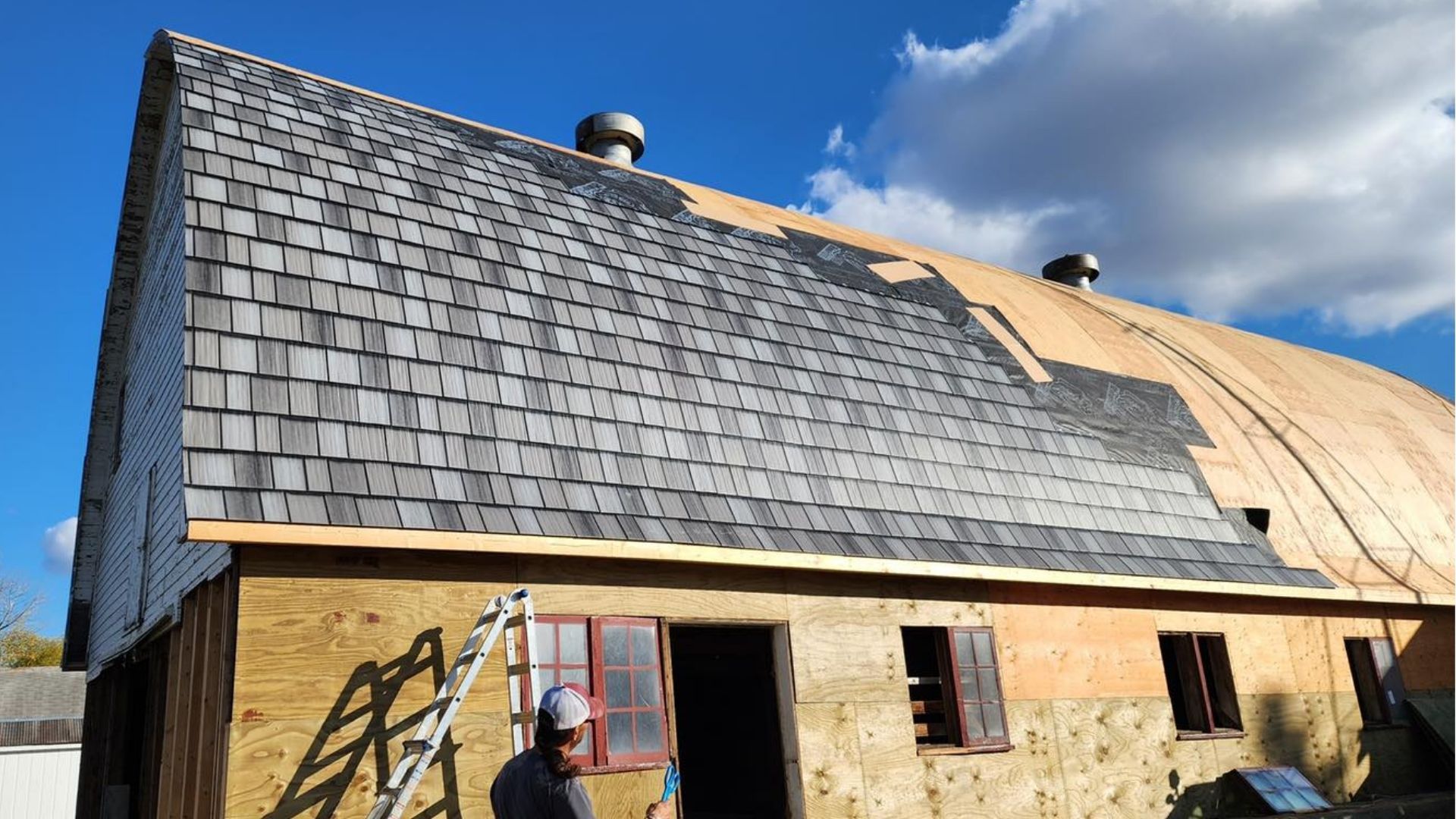 A person works on replacing cedar shake shingles on the roof of a large barn under a sunny blue sky.