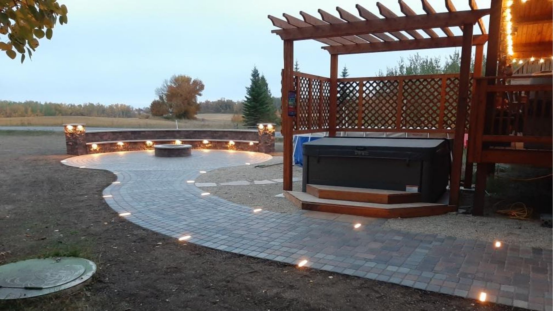 A stone patio with an outdoor hot tub under a wooden pergola, leading to a circular fire pit area with integrated seating.