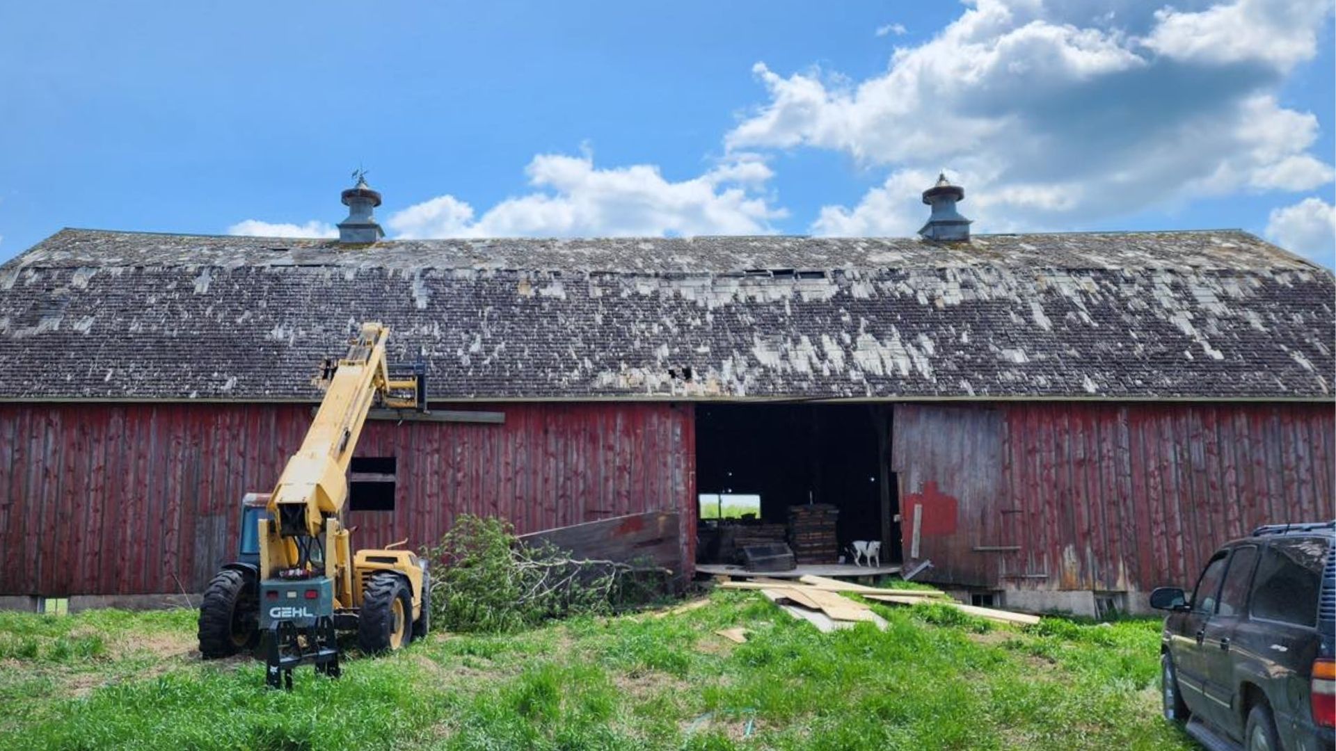 A yellow telehandler sits before a weathered red barn with two cupolas under a blue sky with white clouds.
