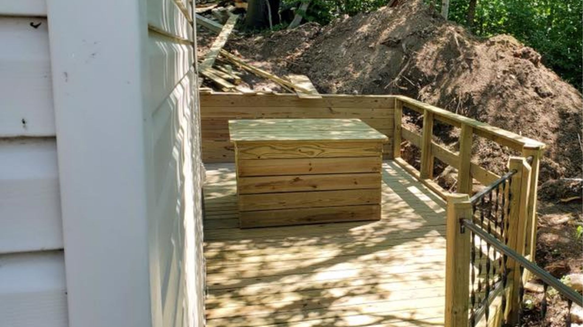 A wooden deck beside a house features a square wooden structure and railings, with a pile of dirt in the background.