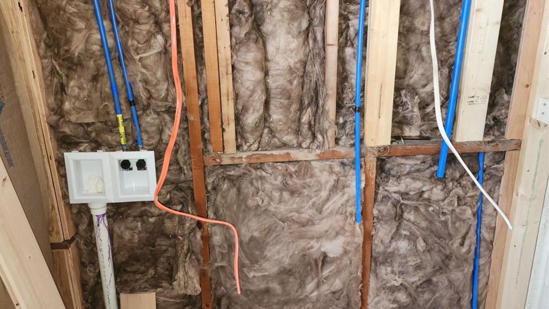 An interior wall under construction, showing wooden studs, fiberglass insulation, blue PEX plumbing pipes, and a white box.