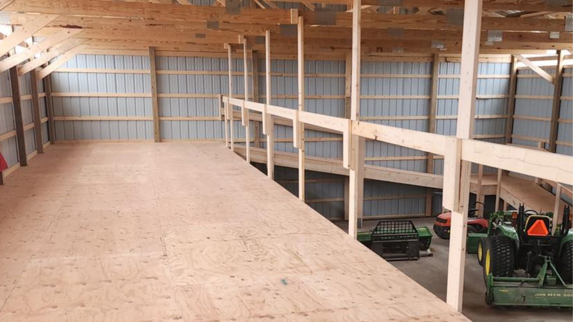 A loft with a wooden floor inside a large barn, overlooking a ground level storage area with farm equipment.