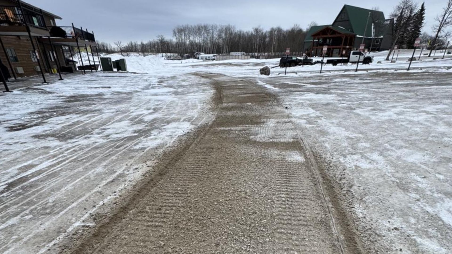 A gravel path, cleared of snow, leads through a snowy, overcast parking lot toward two buildings in the distance.