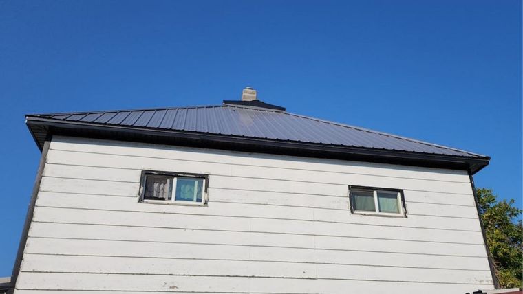 A two-story white house with light gray horizontal siding and a dark metal roof against a bright blue sky.