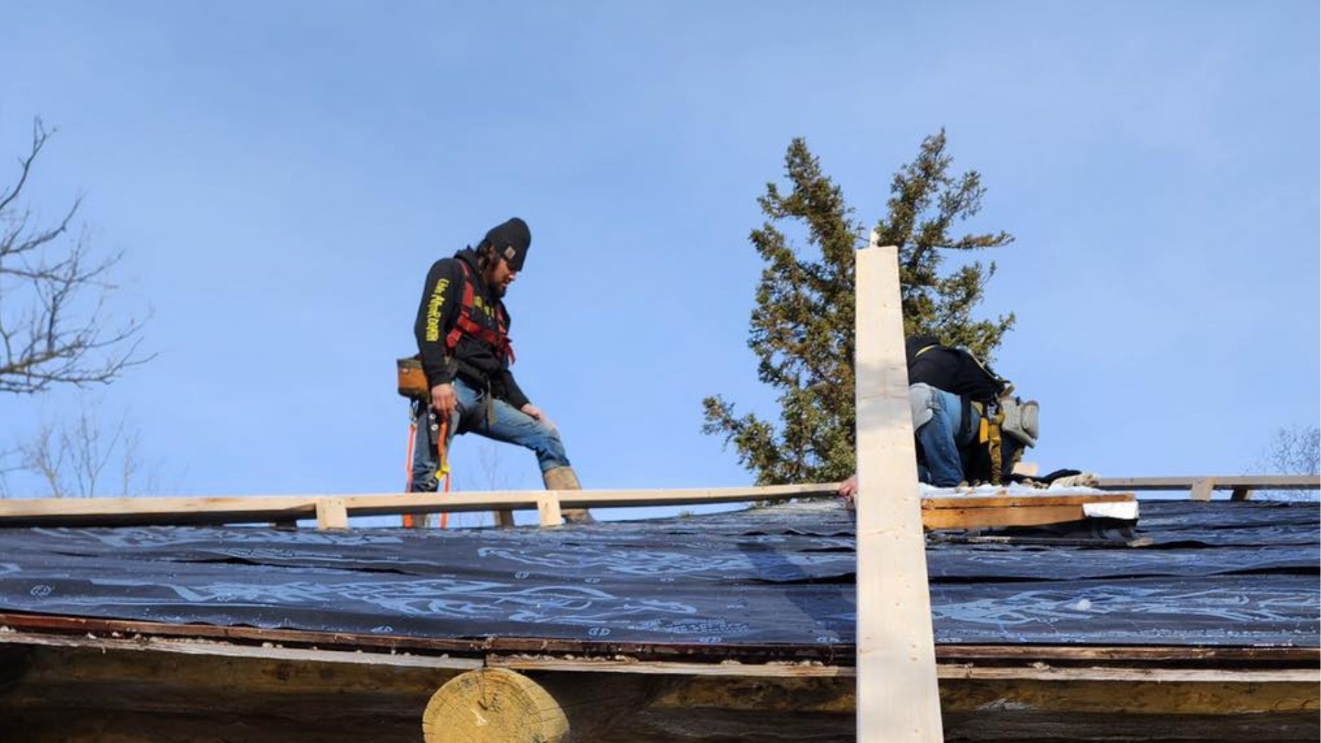 Two construction workers install wooden beams on a slanted roof covered in black underlayment against a blue sky.