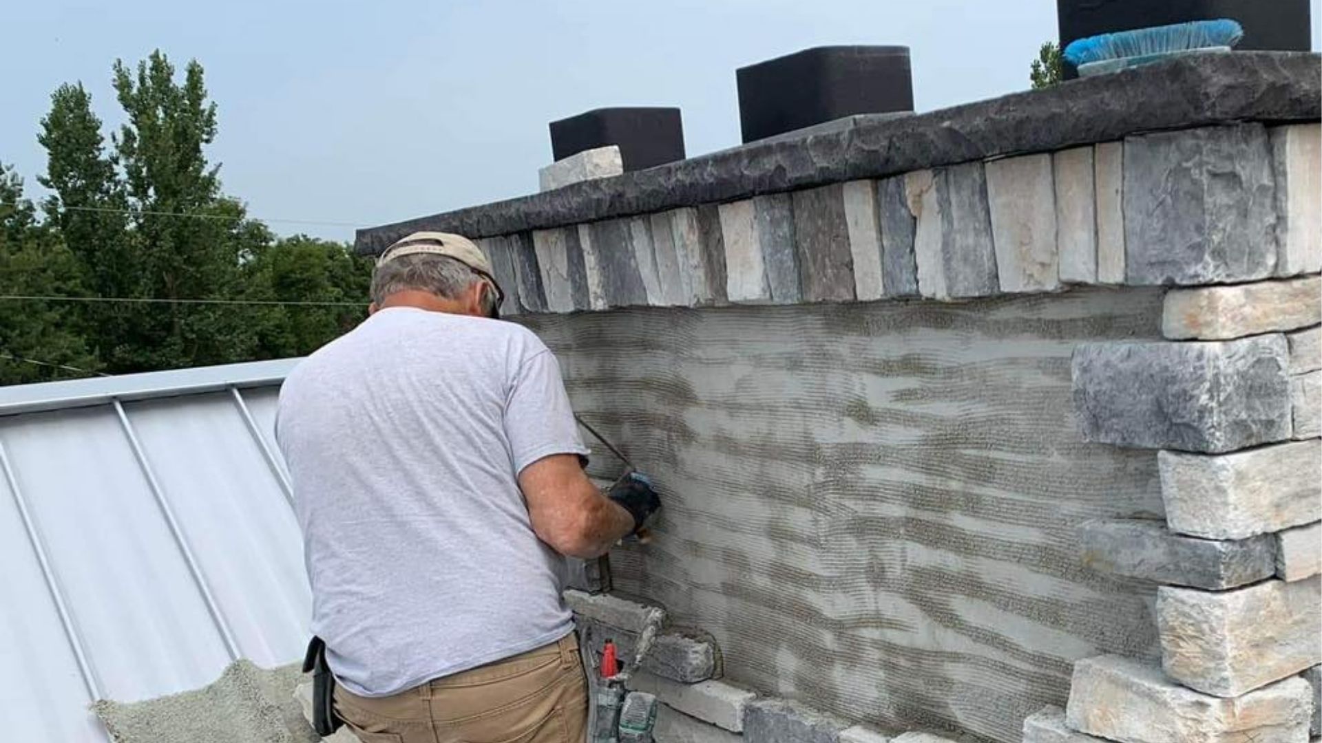 A person in a light-colored shirt works on a stone chimney facade on a metal roof.