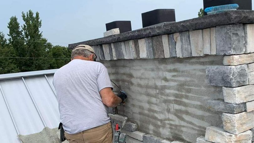 A person wearing a hat works on a stone chimney facade on a roof, applying mortar to the exterior surface.