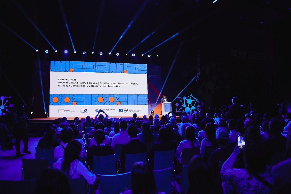 A crowd of people are sitting in front of a large screen at a conference.