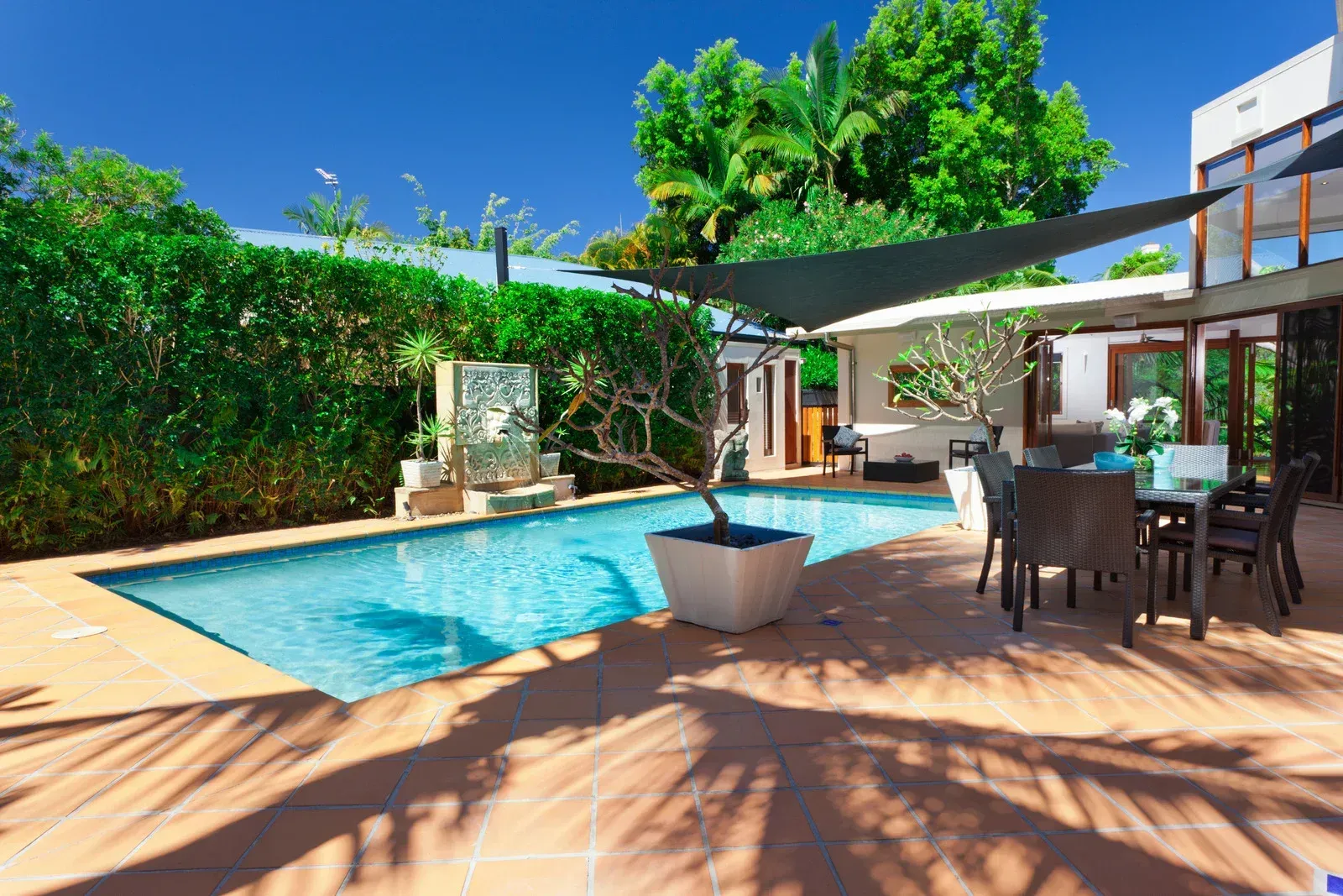 Pool and patio area of a modern home with shade sail, green foliage, and blue sky.