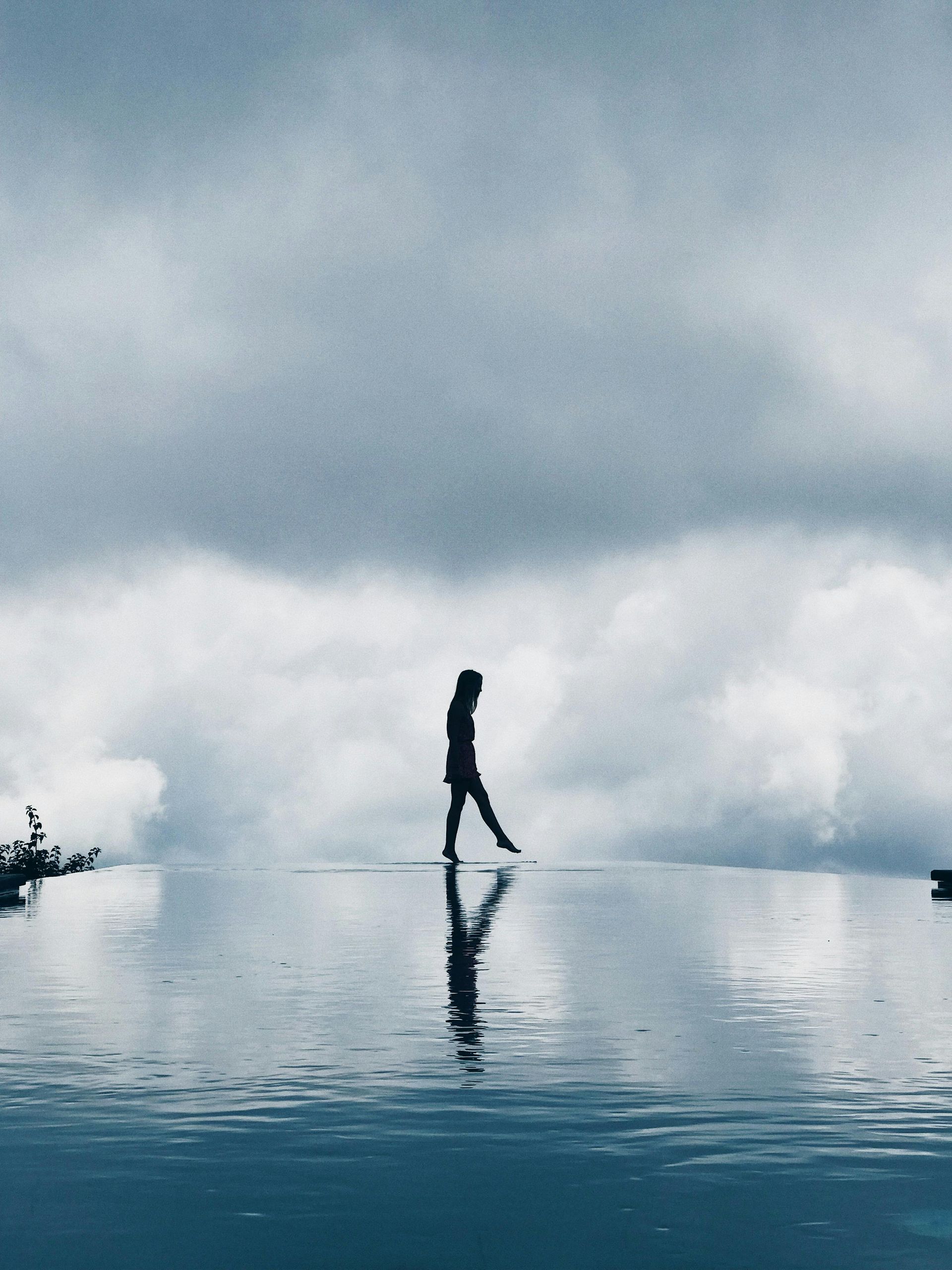 Woman walking on water, reflected in a pool. Cloudy sky above a serene, reflective scene.