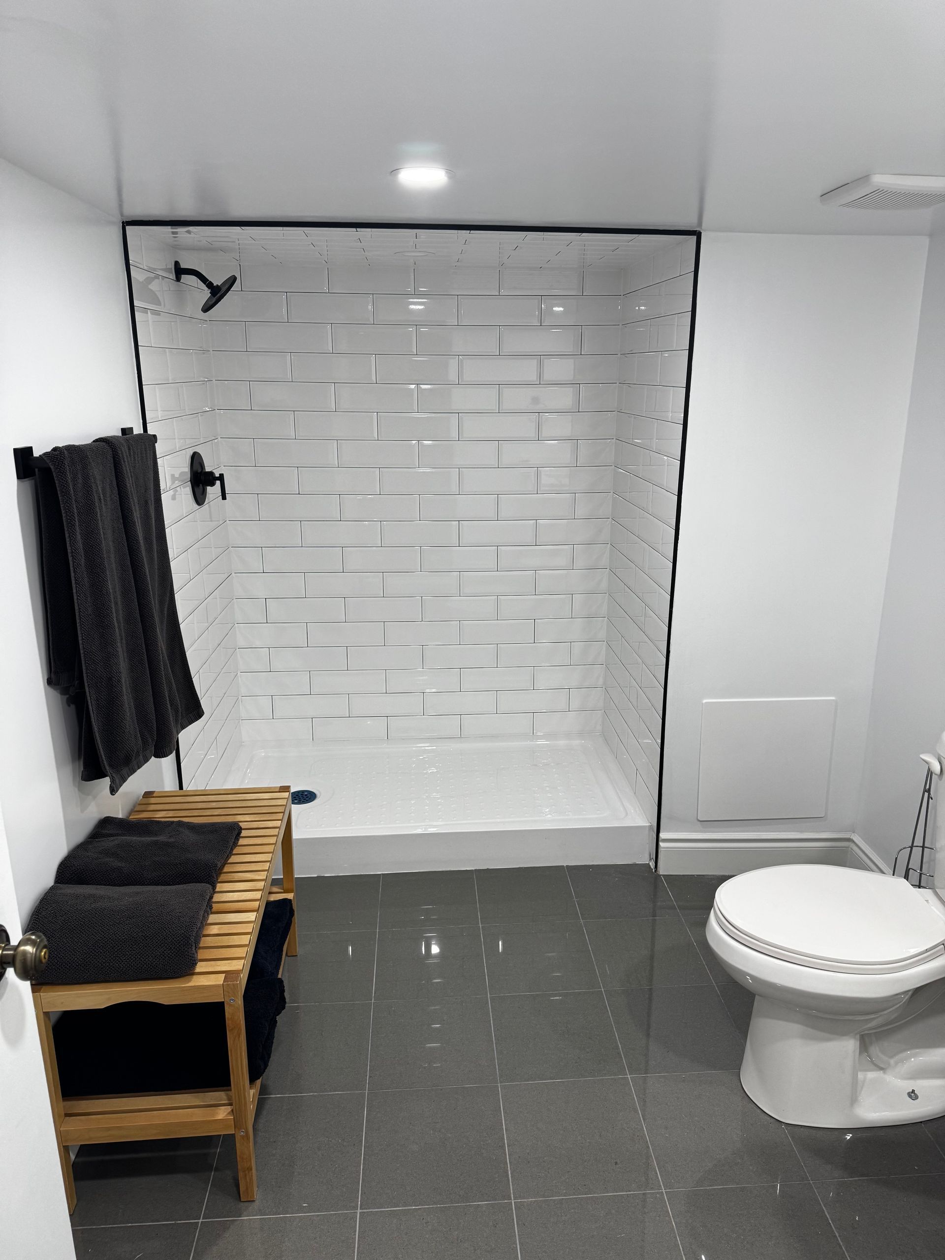 Modern bathroom with white tiled shower, black fixtures, and a wooden bench with towels.