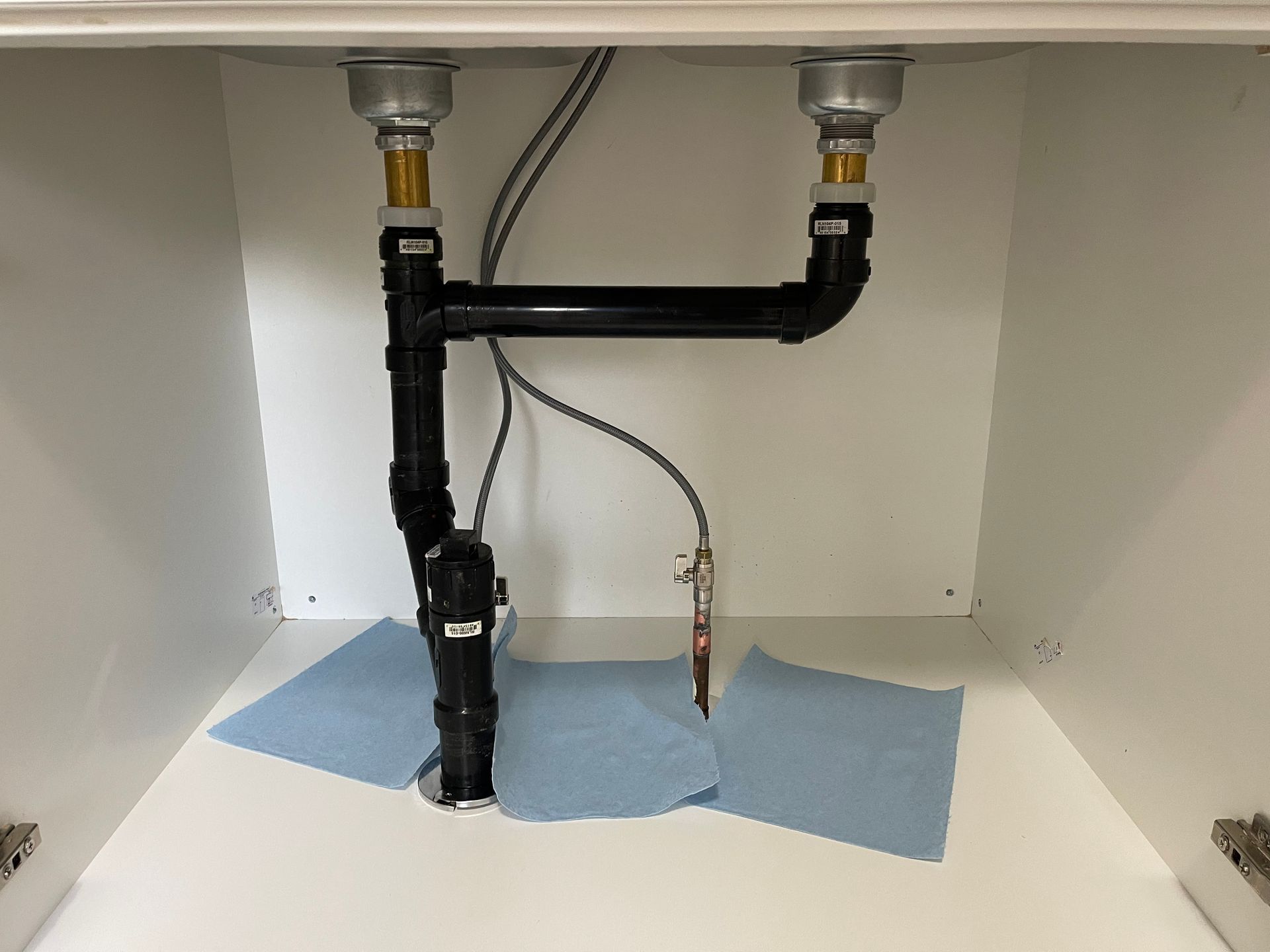 Plumbing under a white sink, with black pipes, chrome fixtures, and blue paper towels.