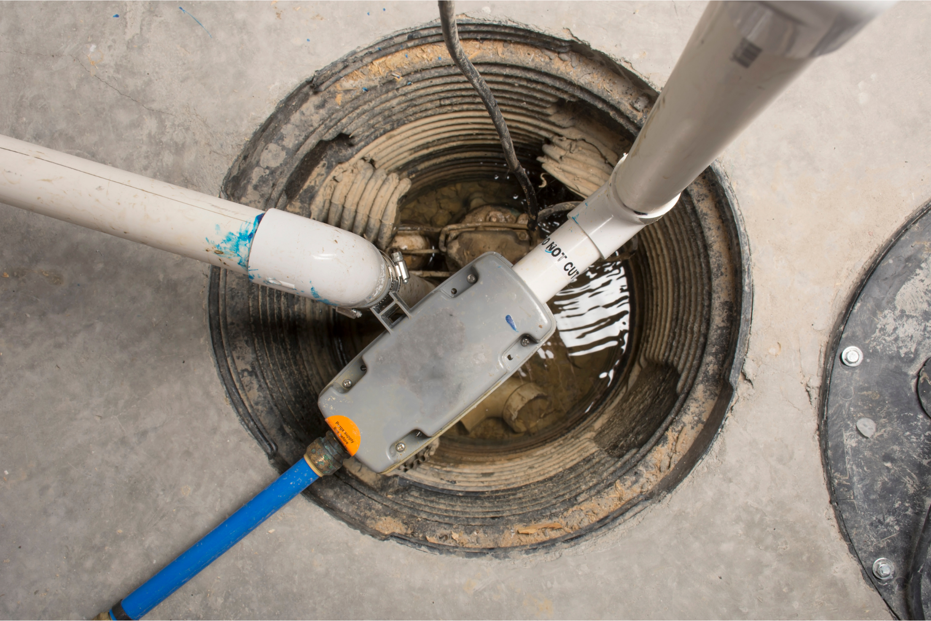 Sump pump pit with water, PVC pipes, and a blue pipe in a concrete floor.
