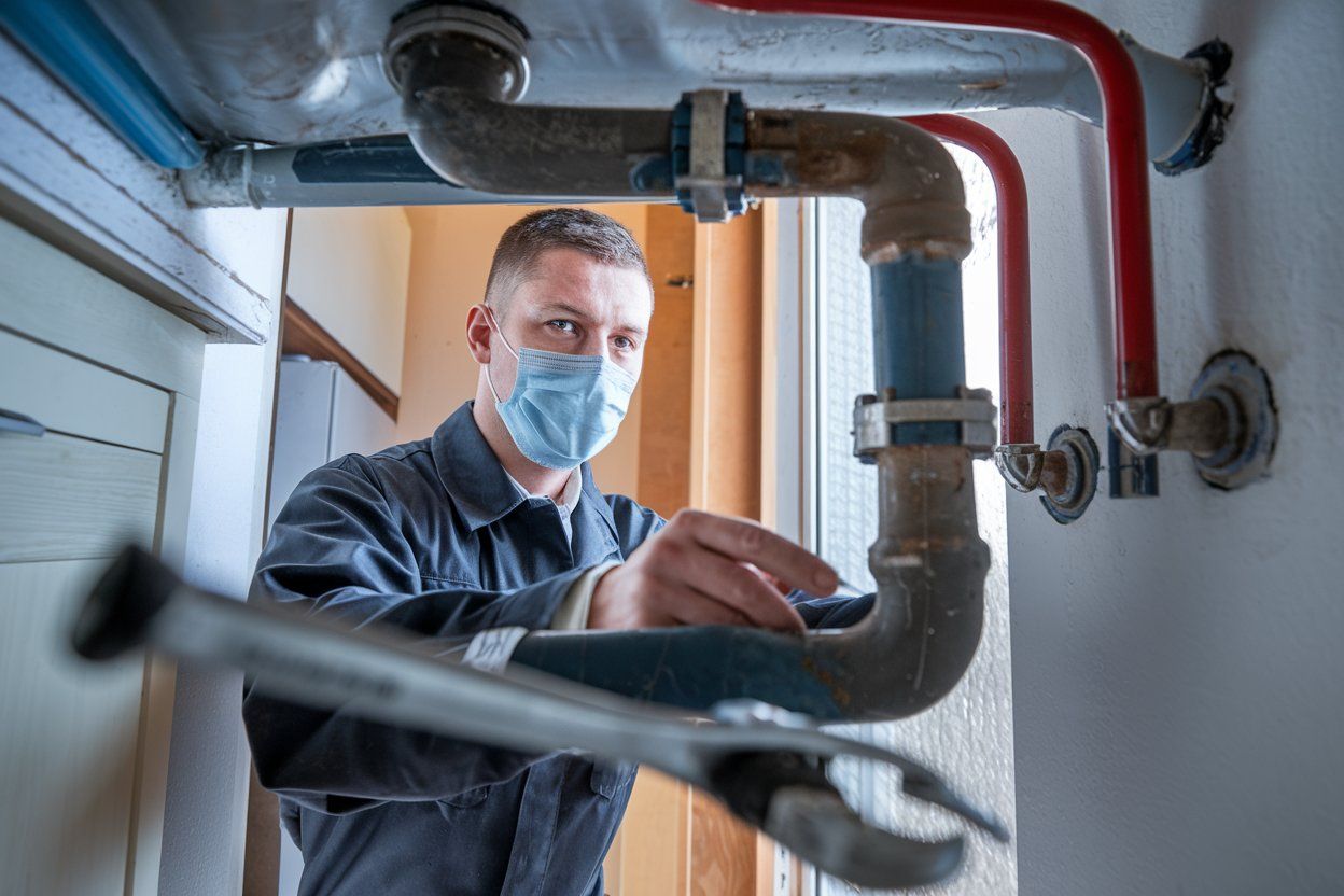 Plumber in face mask using a wrench on pipes under a sink.