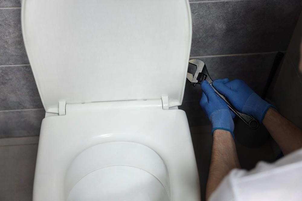 Person with blue gloves uses a wrench to tighten a toilet seat hinge. White toilet, gray tiled wall.