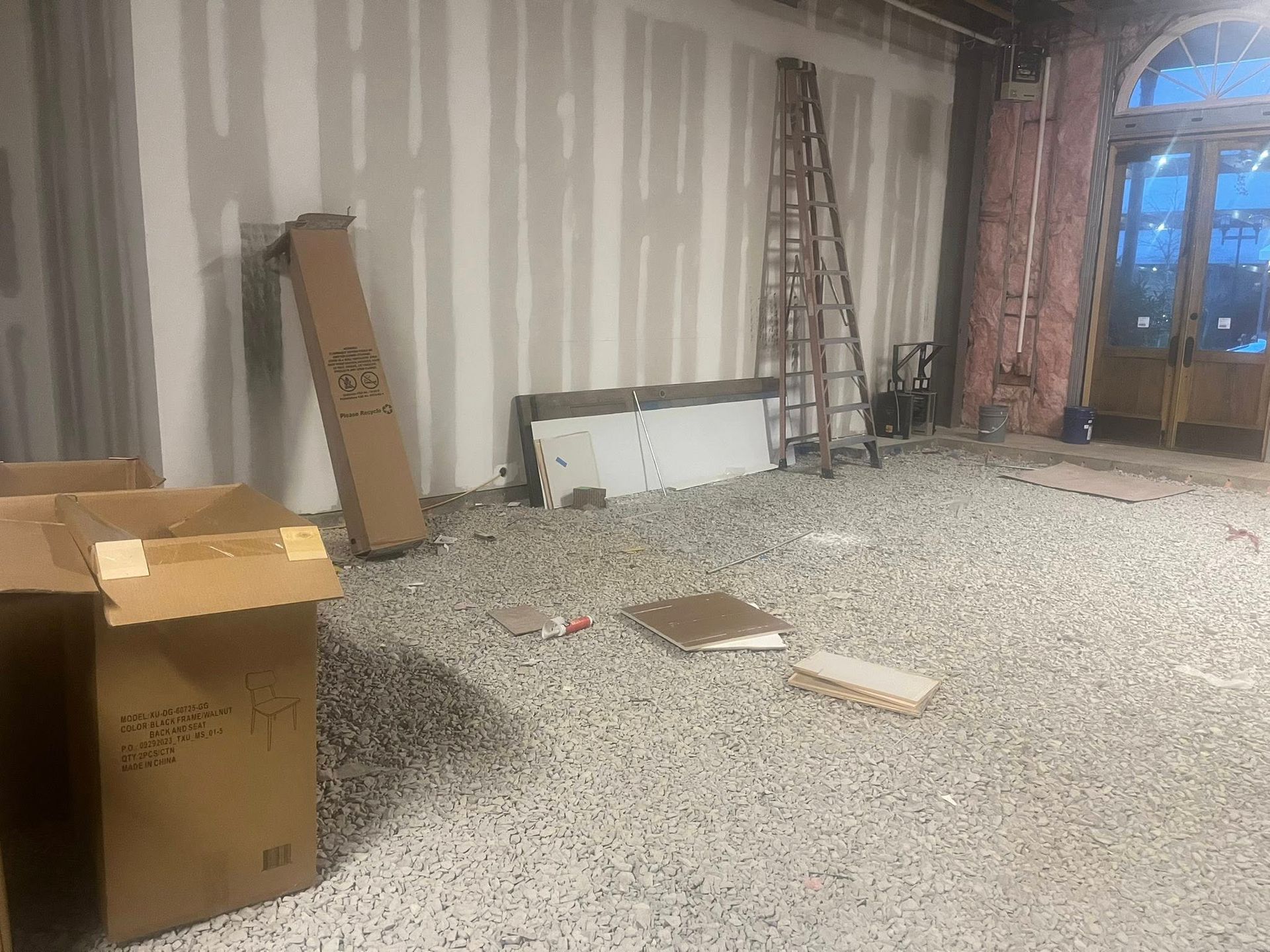 Indoor construction site featuring a gravel floor, taped drywall, a ladder, cardboard boxes, and an arched doorway.