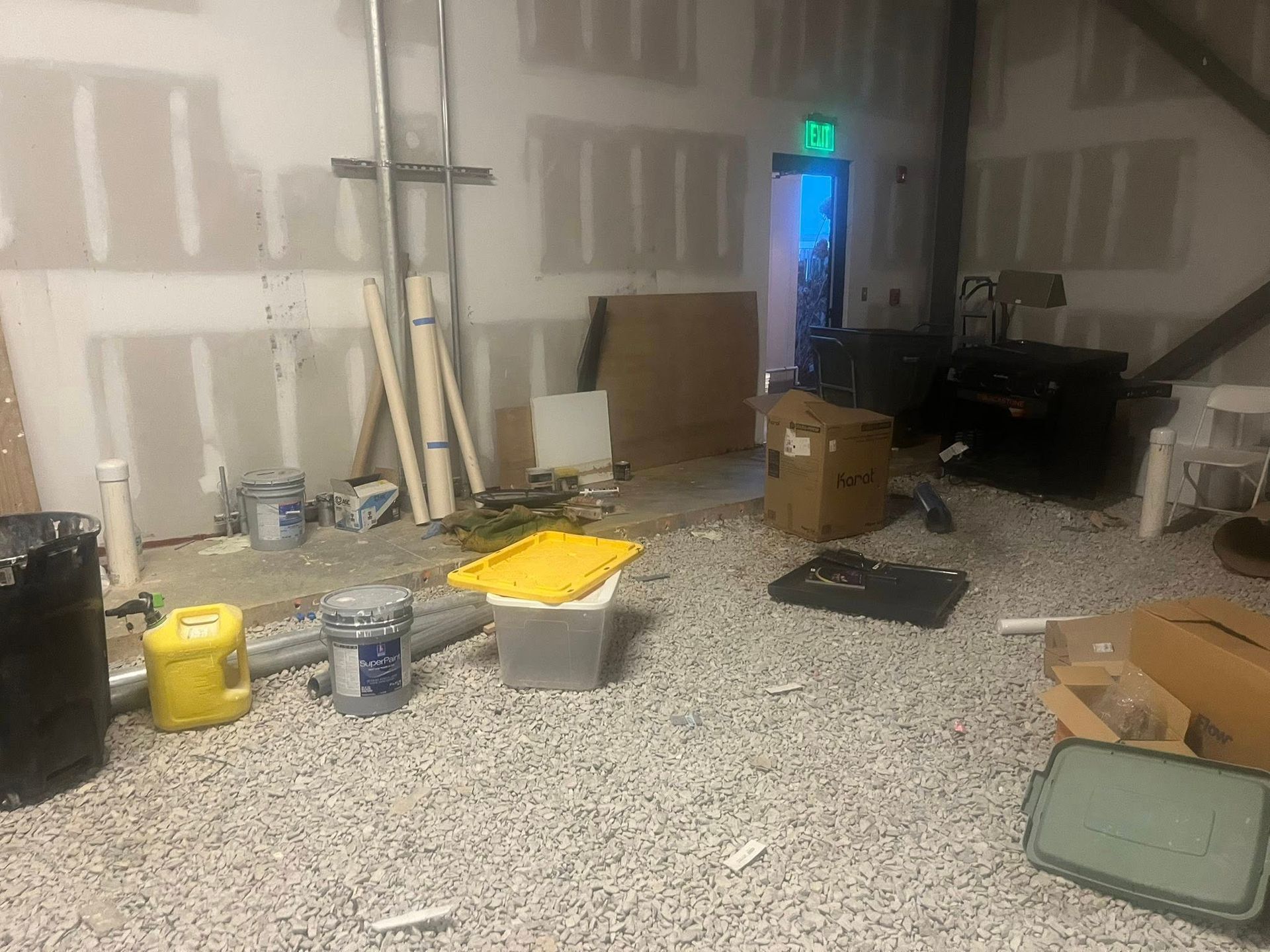 A cluttered room under construction with white drywall, a speckled floor, and various boxes, bins, and building materials.