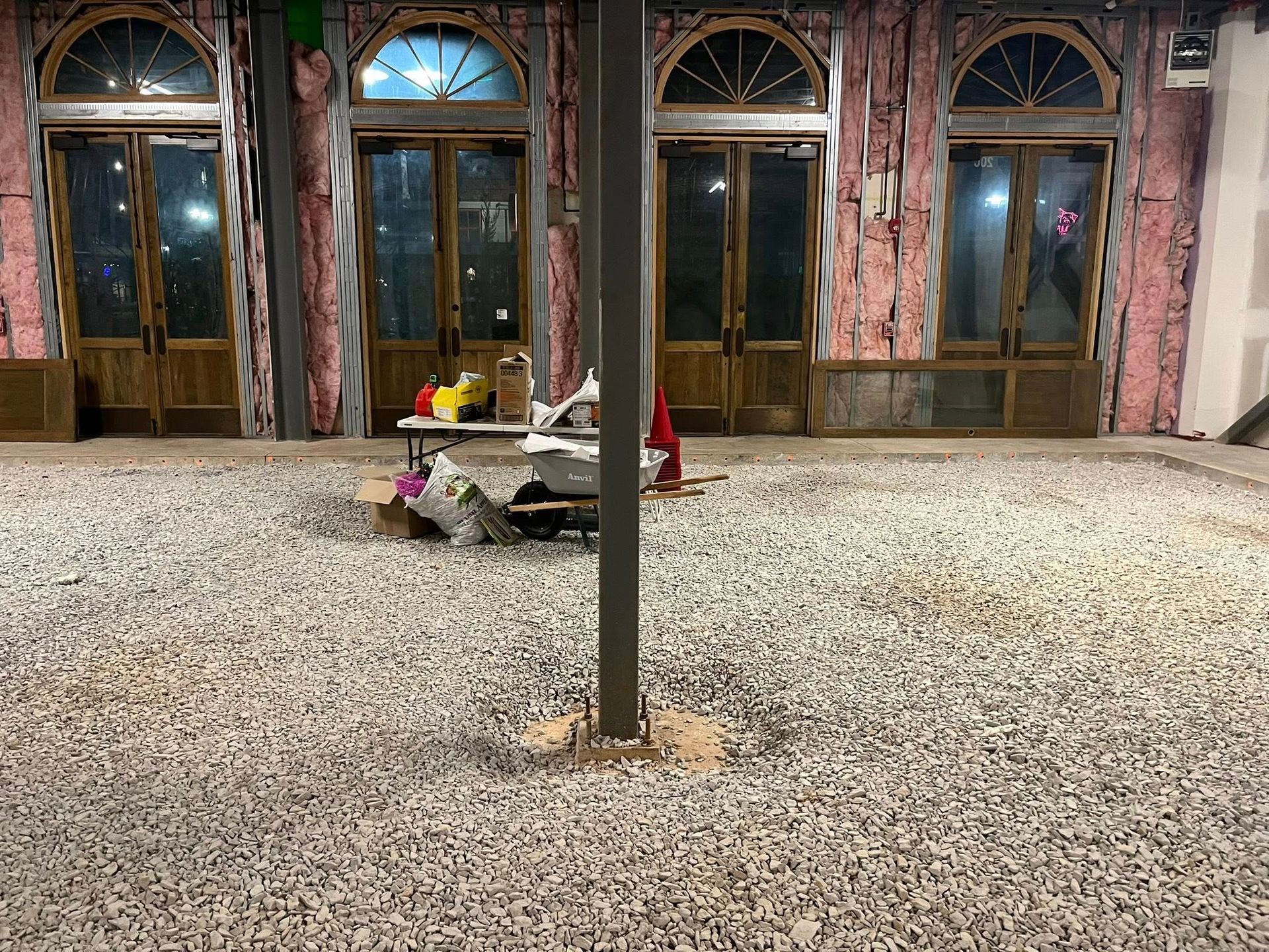 Interior of a room under construction with a gravel floor, a central support column, and four arched wooden glass doors.