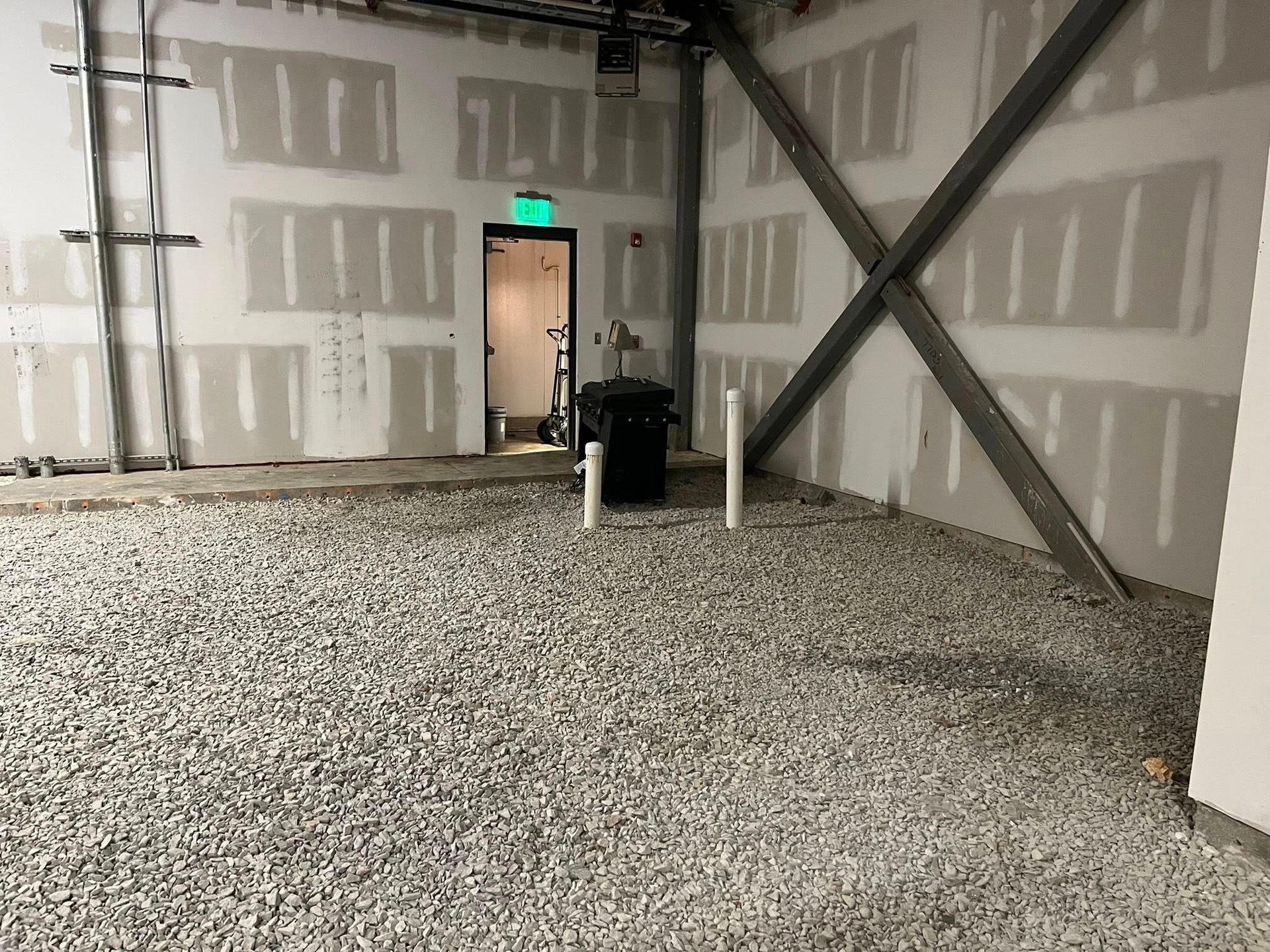 Unfinished interior space with a gravel floor, metal X-bracing on the wall, and an open doorway with an exit sign.
