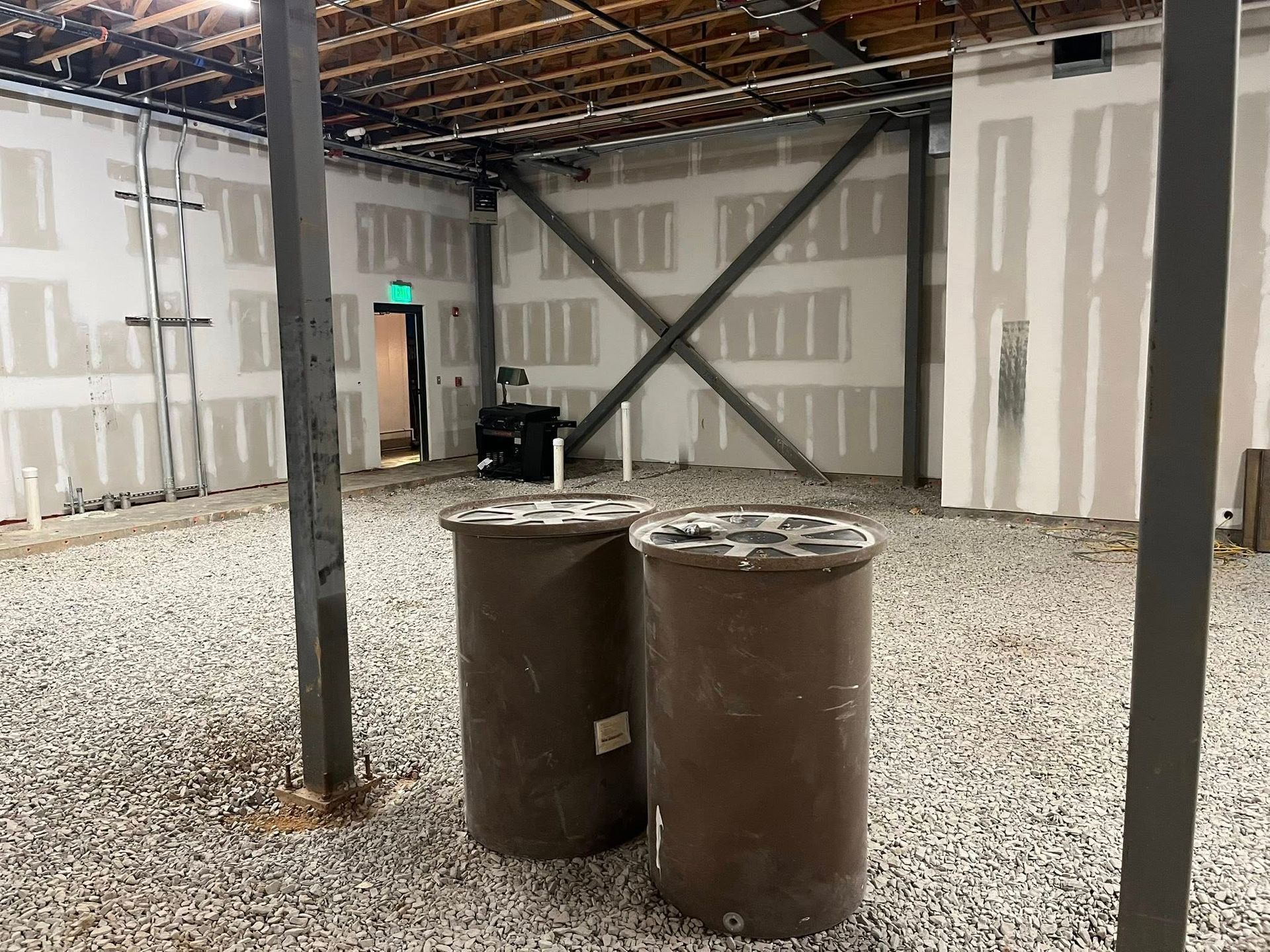 An unfinished interior space with gravel flooring, exposed ceiling beams, drywall walls with X-bracing, and two large barrels.