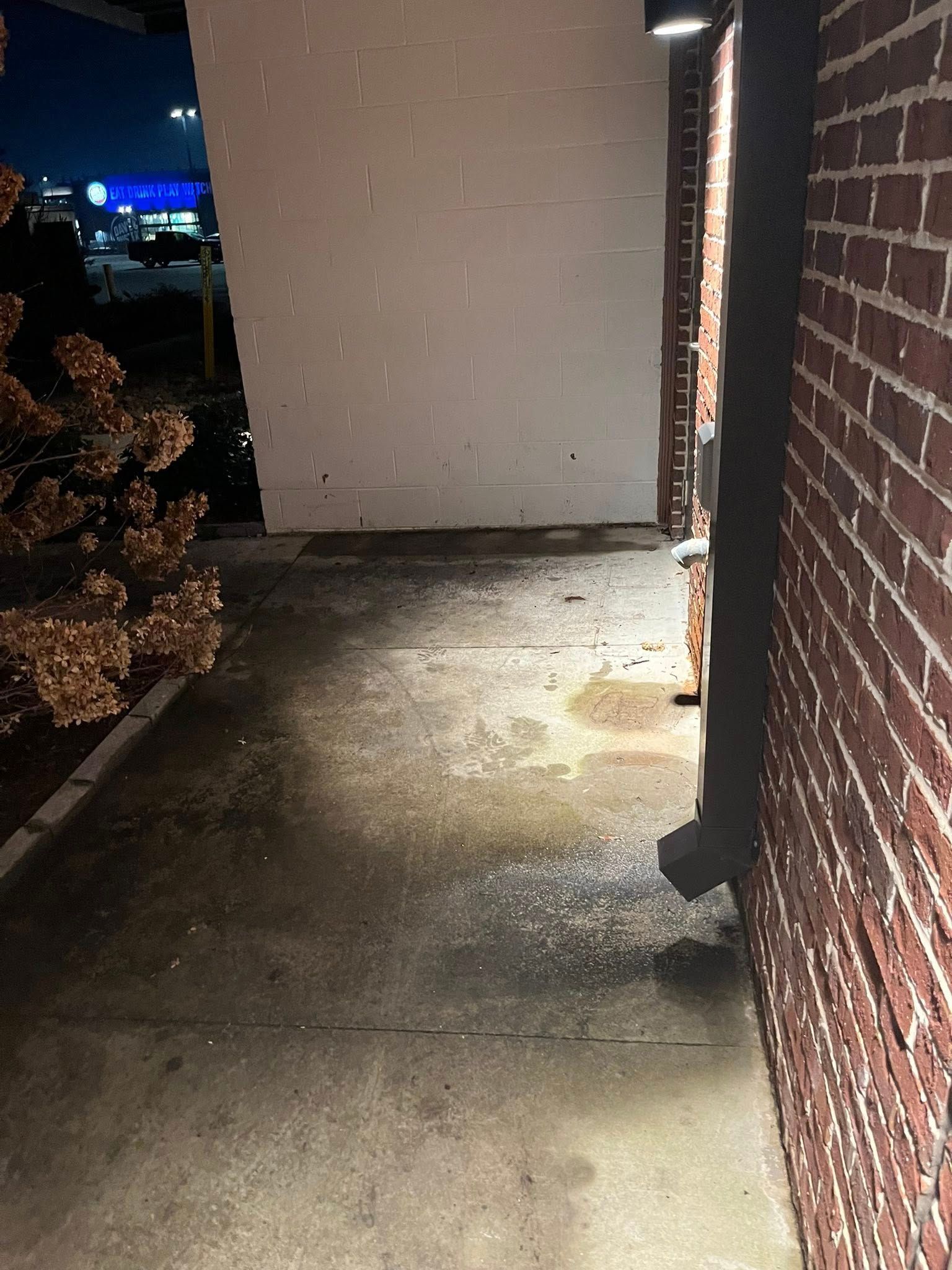 A lit concrete walkway at night runs between a white wall and a red brick wall with a dark downspout.
