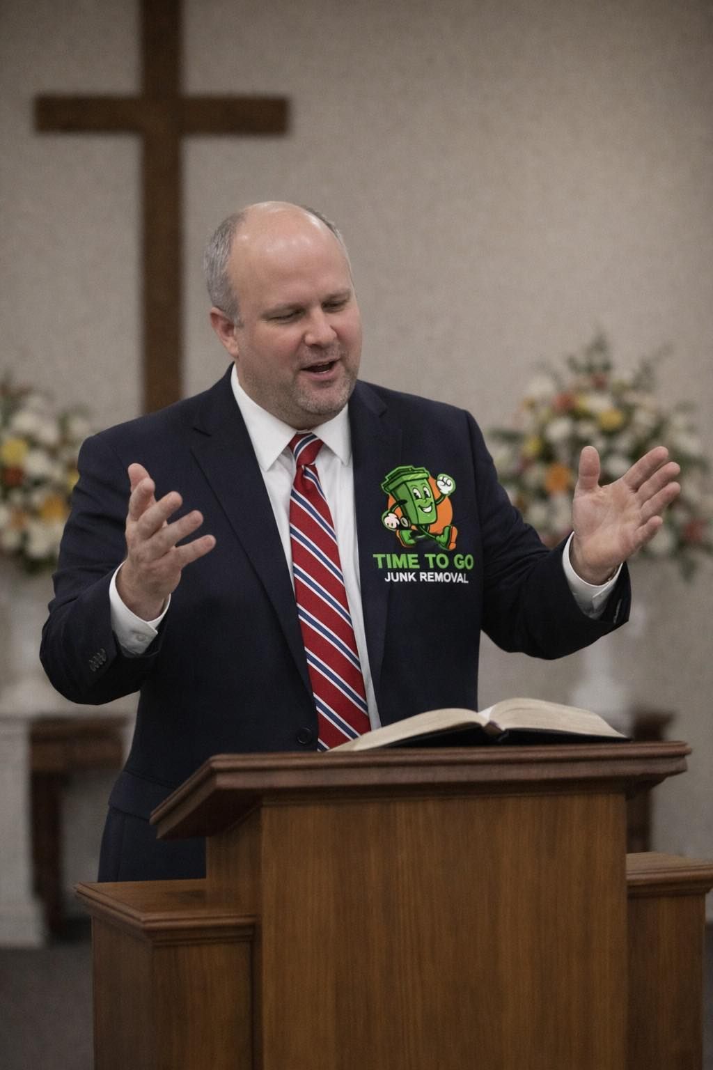 A speaker in a suit and tie gestures with open hands while standing at a wooden lectern in front of a cross.