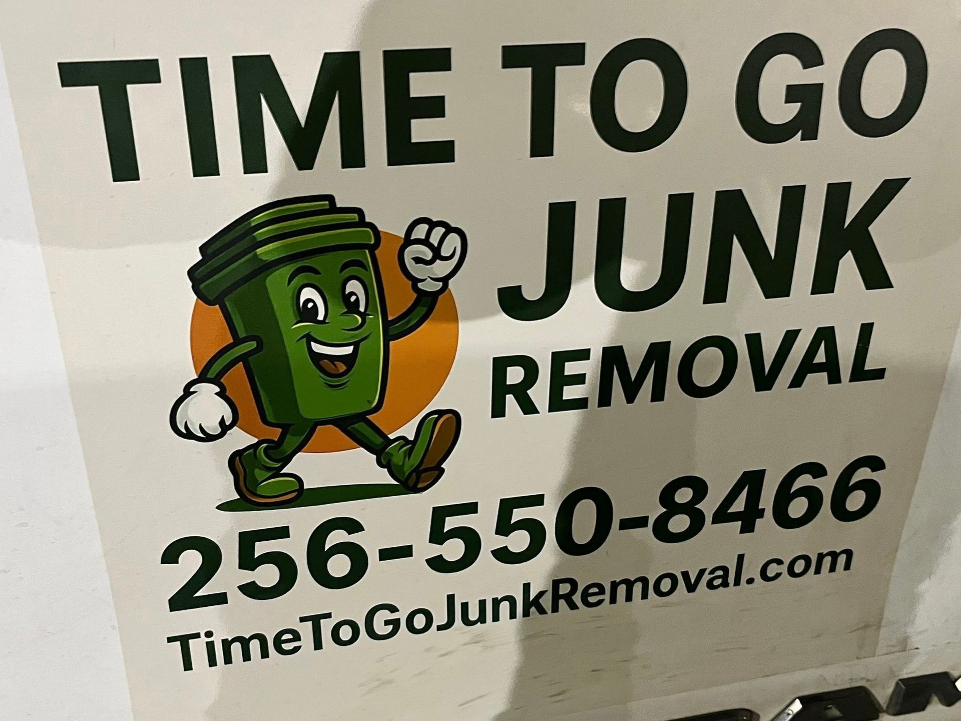 Sign for Time To Go Junk Removal featuring a cartoon green trash bin, phone number 256-550-8466, and website address.