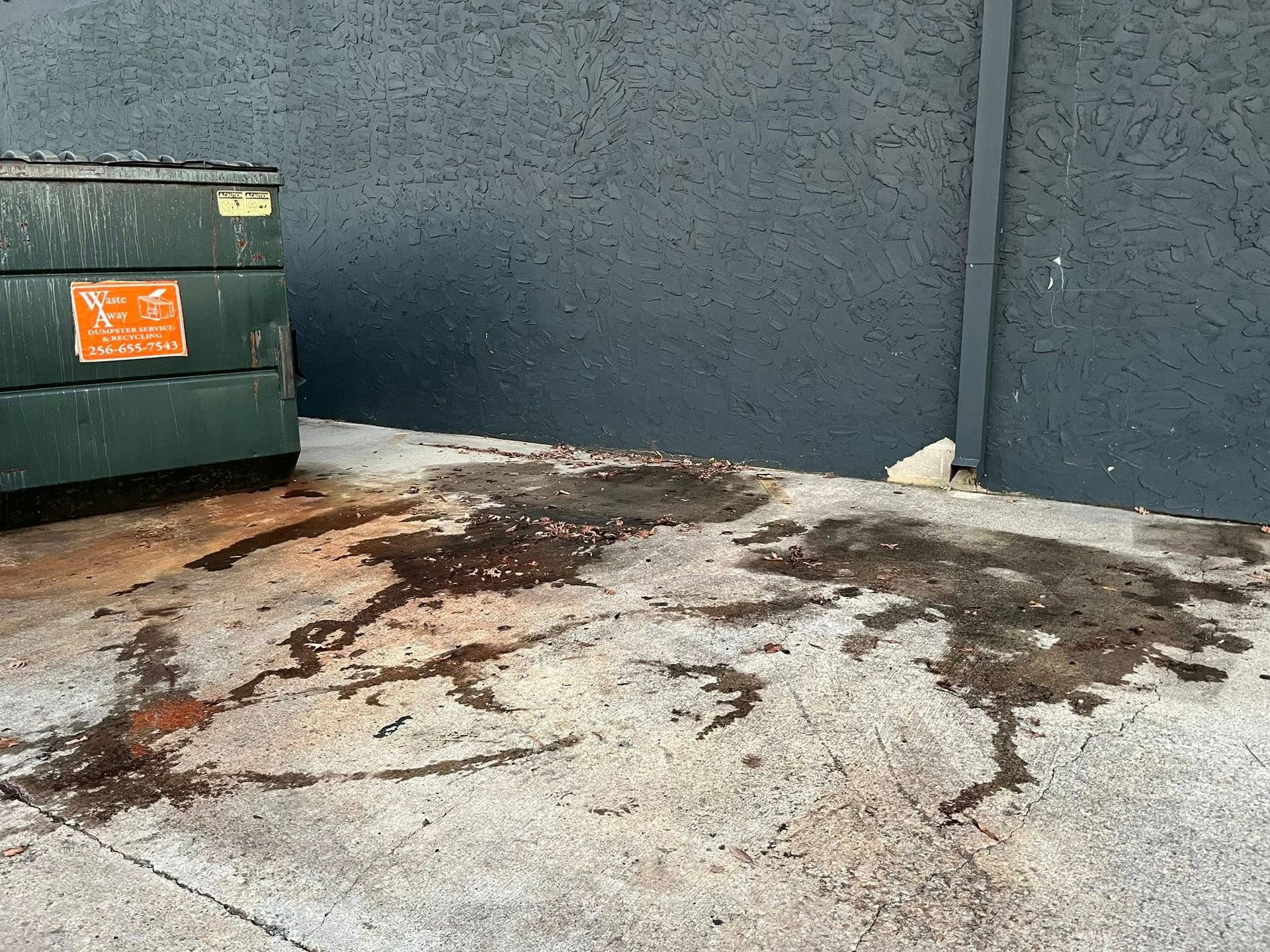 A green dumpster sits next to a dark gray wall on a concrete surface stained with dark, irregular patches of liquid.