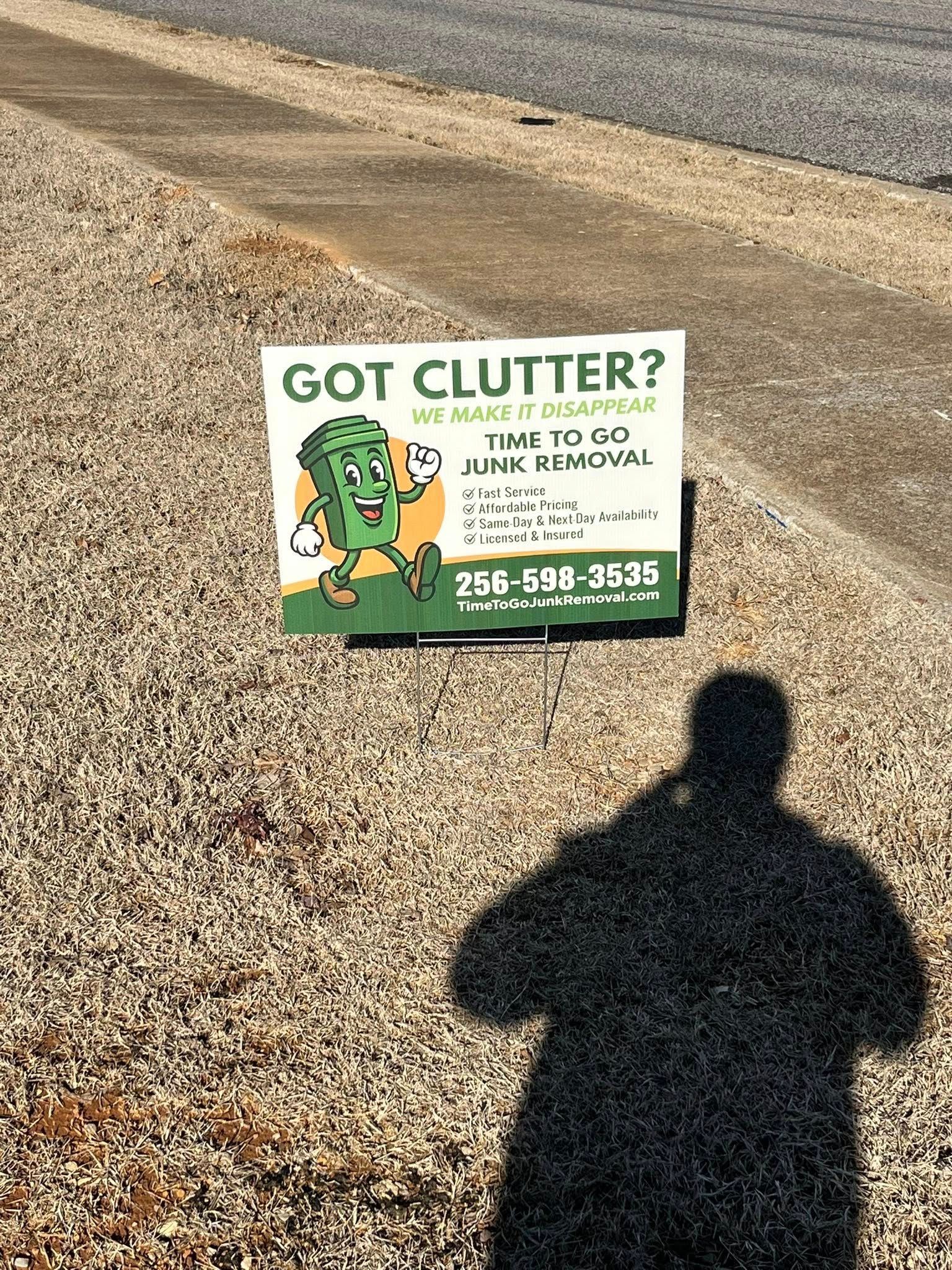 A yard sign for junk removal services with a green cartoon trash can character, phone number 256-598-3535, on gravel.