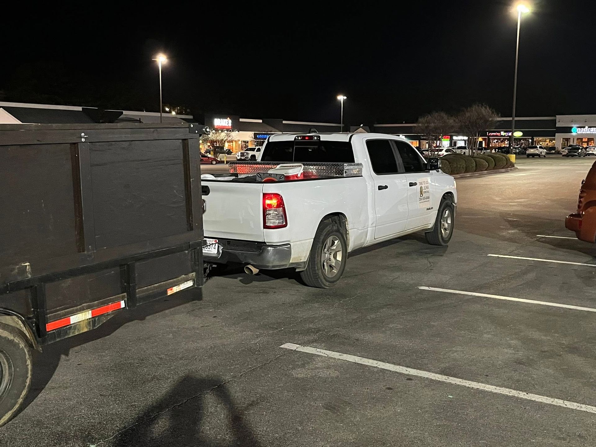 A white pickup truck with a large dark trailer attached is parked in a retail parking lot at night.